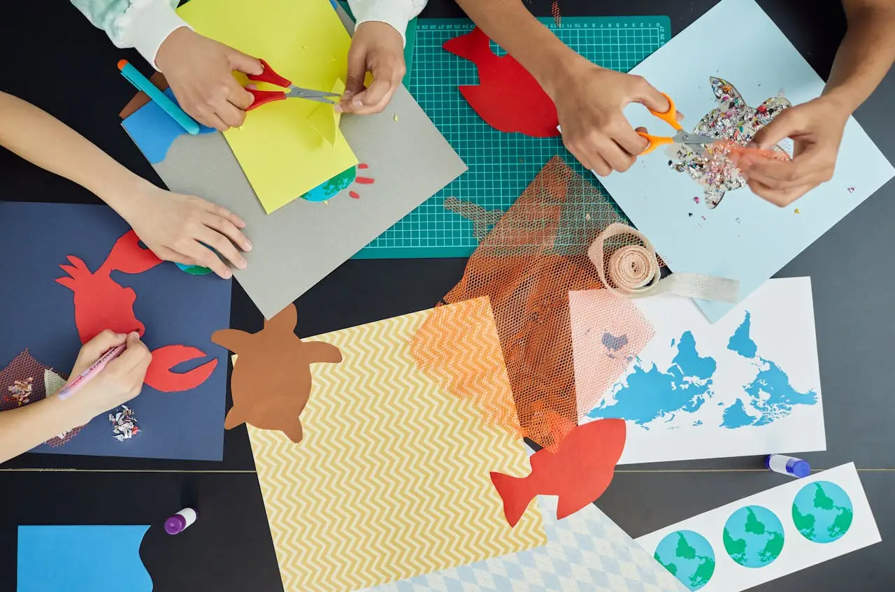 Top view of group of crop anonymous children sitting at table and creating colorful draft and artworks with paper and scissors